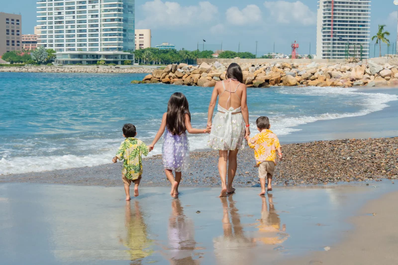 A mother and three children walking away along the shoreline, holding hands, reflections in the wet sand.